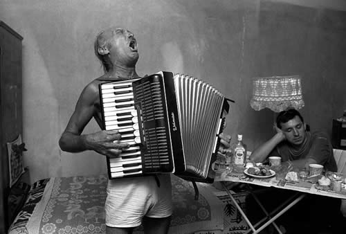 B&W photo of a man wearing only shorts playing accordion in a living room.
