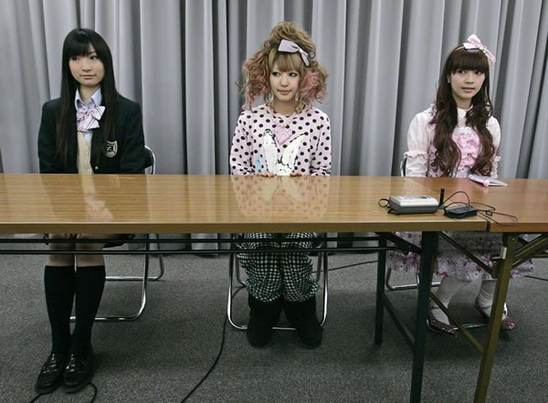 Three young Japanese women at a press conference table, one dressed in a schoolgirl outfit, one in a "Harajuku" outfit and one in a "Lolita" outfit.