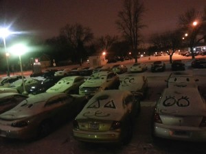 Snow-covered cars in a parking lot, with faces drawn on their windshields