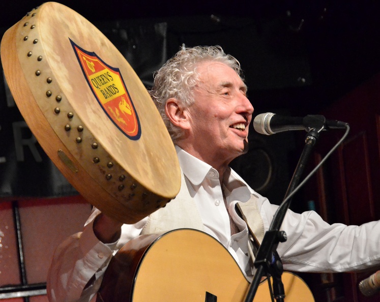 Gerry O’Kane onstage with guitar, holding up a bodhran (Irish hand drum) with the Queen’s University Bands logo painted on it