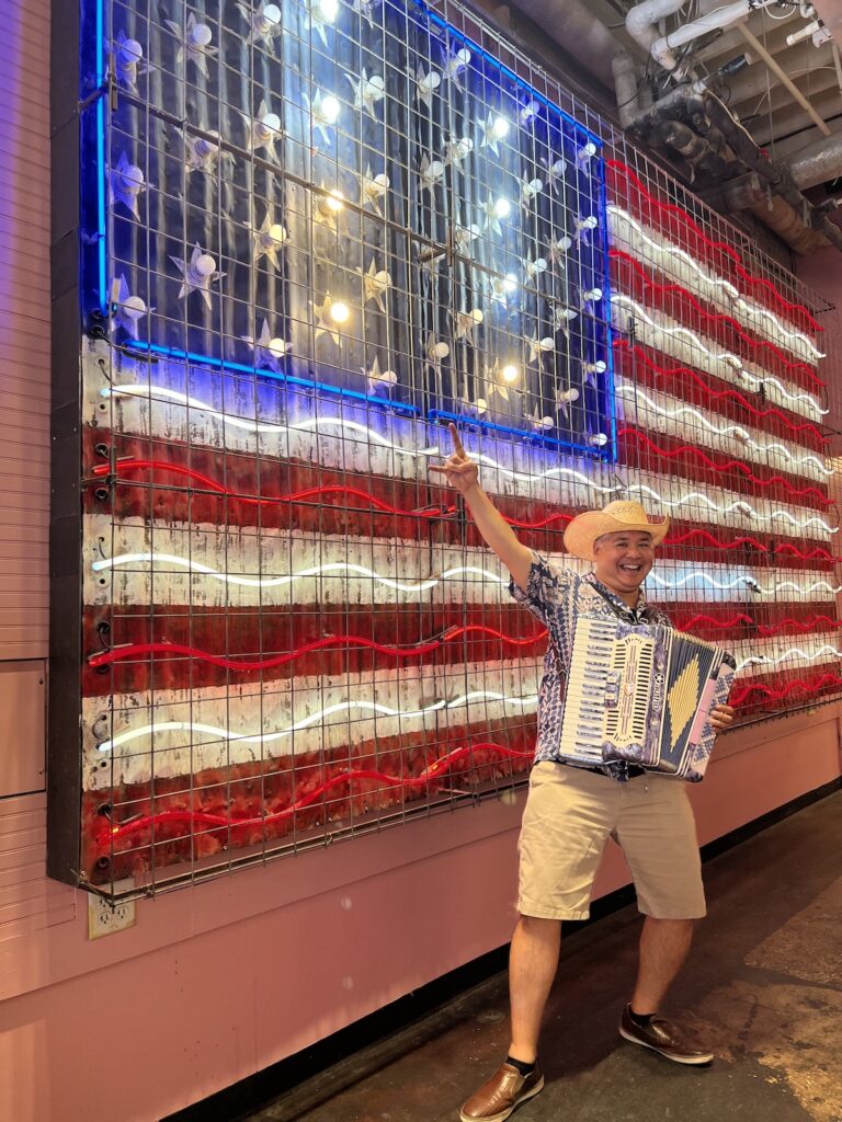 Joey deVilla posing with accordion and cowboy hat in front of the American Flag ighting display at Voodoo Donut in Austin.
