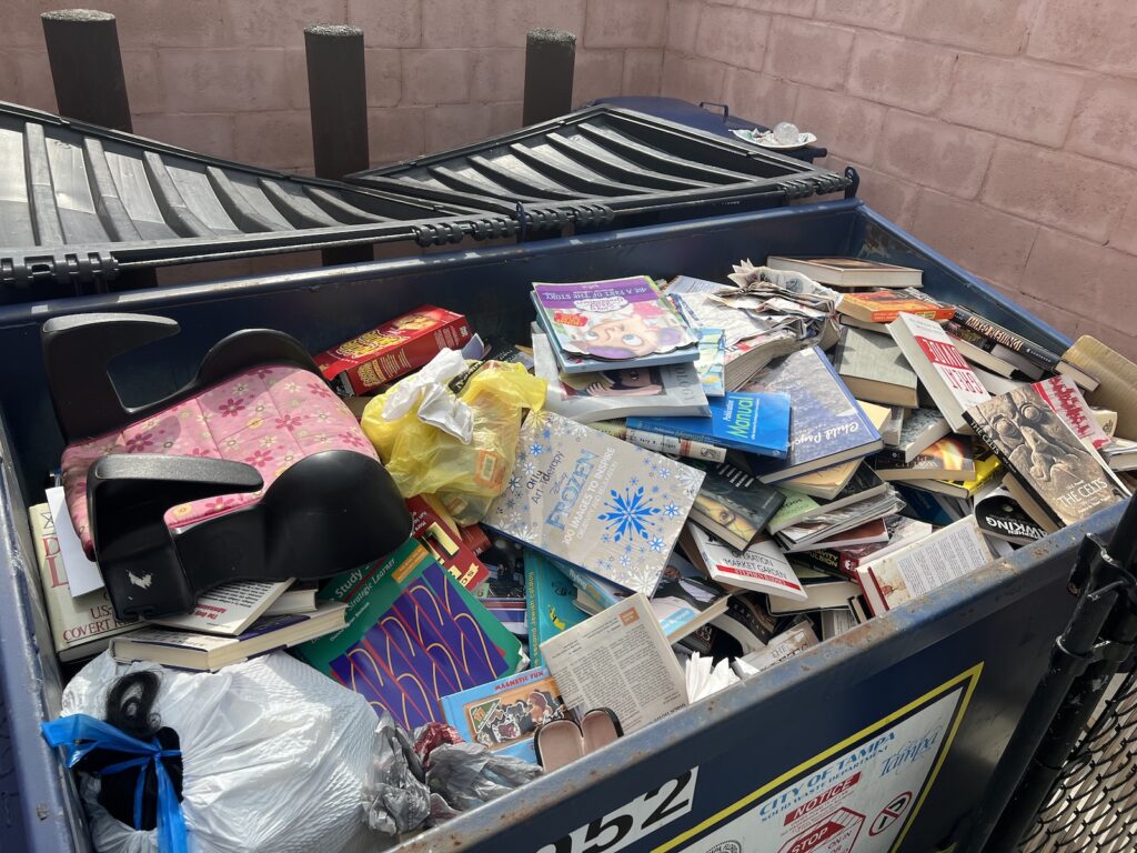 The dumpster, filled with books, along with a discard child’s car seat, and some regular garbage.