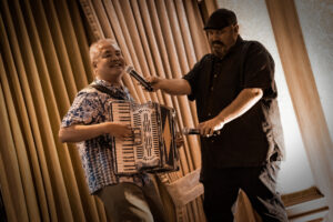 Joey deVilla plays accordion while a karaoke DJ points two microphones at him