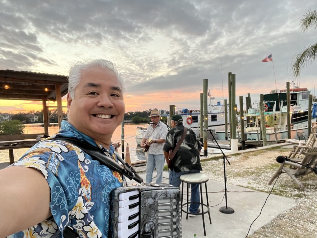A selfie featuring Joey deVilla in an aloha shirt with his accordion in the foreground, and Tom Hood and the Tropical Suns in the background. Behind them is Tarpon Bay and a Florida sunset.