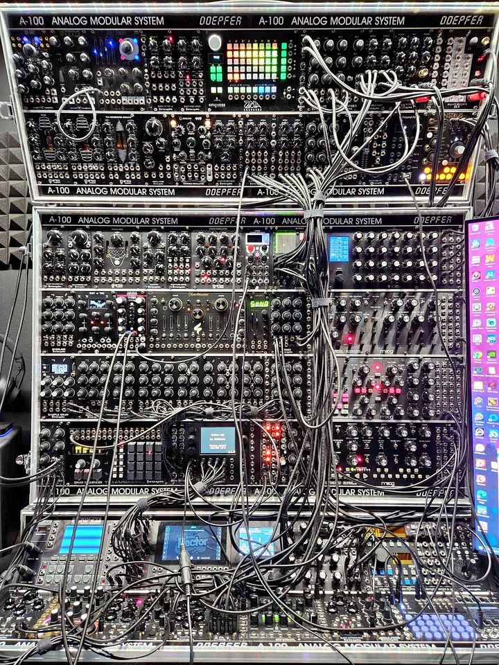 A massive setup of various analog synthesizer modules — think of a commercial airplane cockpit, but with more patch cables — in three large racks, effectively creating a wall of knobs, buttons, cables, and the occasional LCD screen.