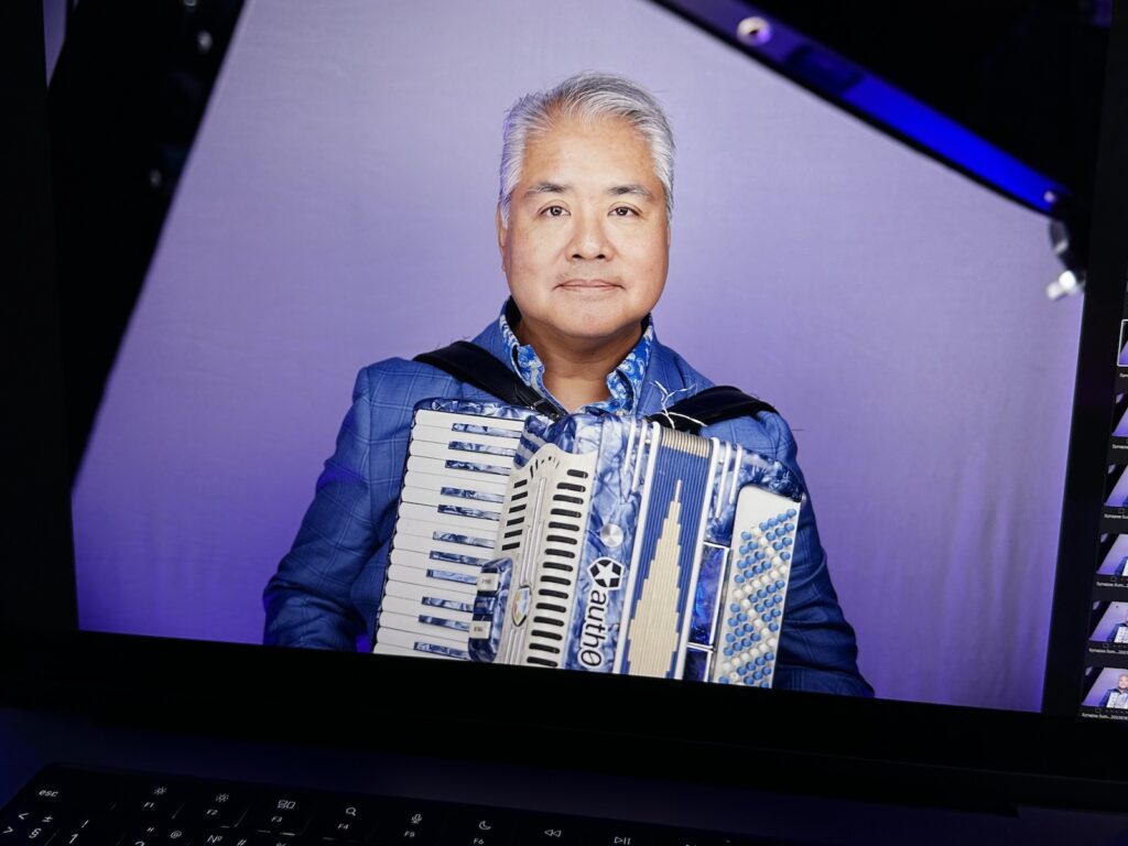 Joey deVilla’s new headshot, showing him in a blue suit jacket and blue shirt holding his blue accordion, as seen from a MacBook.