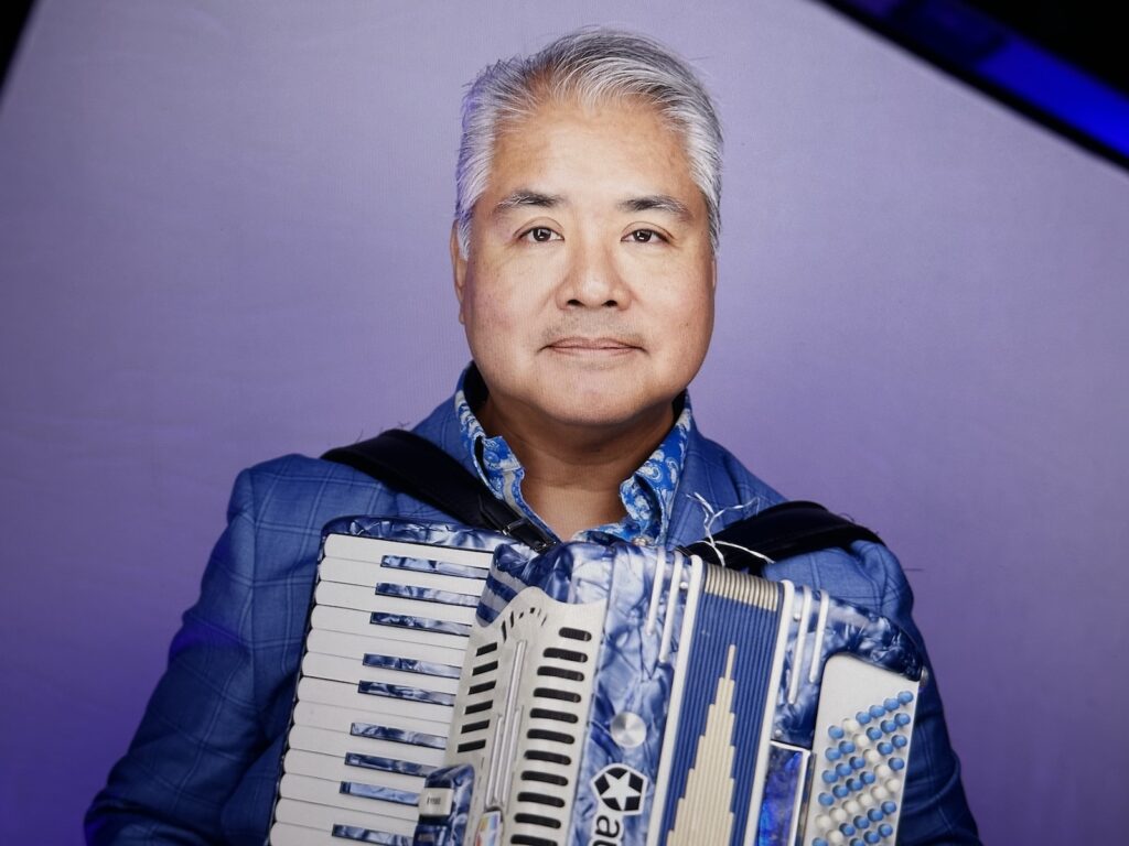 Joey deVilla’s new headshot, showing him in a blue suit jacket and blue shirt holding his blue accordion, as seen from a large display.