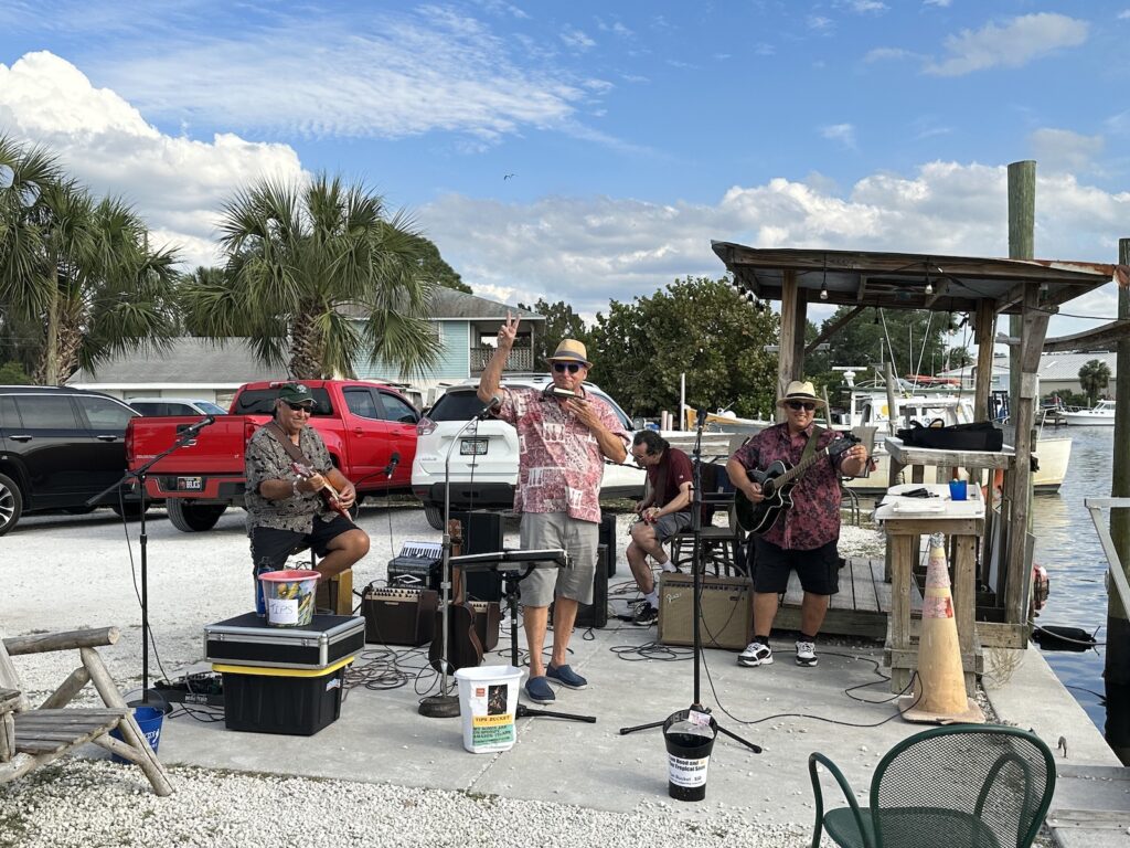 The band “Tom Hood and the Tropical Sons,” as seen from the audience. In the background is Bayou Bistro’s parking lot, palm trees, and a little bit of Tarpon Bayou.