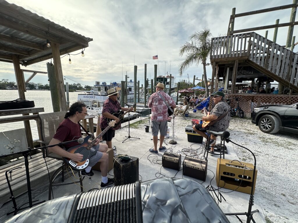 The band “Tom Hood and the Tropical Sons,” as seen from Joey’s vantage point upstage, with Joey’s accordion in the foreground. Pictured from left to right are Richie playing bass, Jay on acoustic guitar, Tom Hood on ukulele, and Dave on electric guitar. In the background are a dock, fishing boats, and Tarpon Bayou.