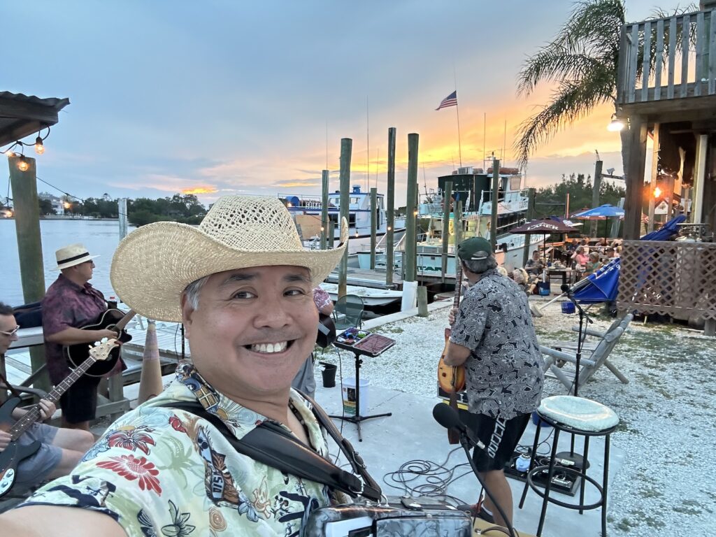 Joey deVilla and accordion in the foreground, with Tom Hood and the Tropical Sons behind him, playing their instruments. In the background are a sunset, a dock, fishing boats, and Tarpon Bayou.