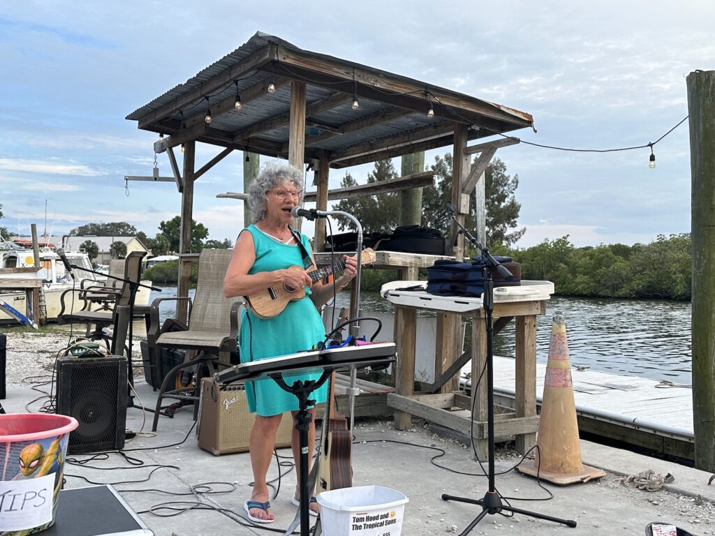 Mary plays her brand new ukulele solo. In the background is the fishing shed, a boat, and Tarpon Bayou.