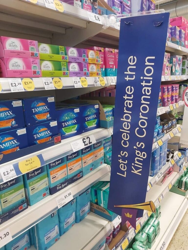 The Tampax shelves in a UK drugstore aisle, marked with a sign that reads “Let’s celebrate the King’s coronation.”