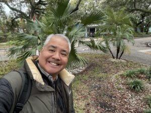Joey de Villa in a swetaer, wool blazer, and puffy vest, smiling in his front yeard, with palm trees behind him.